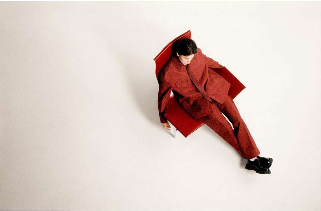 Overhead shot of a man in a patterned red suit lounging on a red chair against a bright white backdrop in a fashion editorial style.