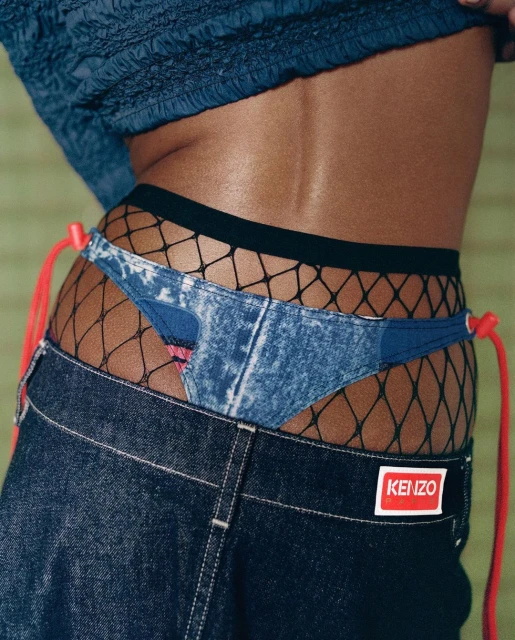 Torso shot of model wearing denim jeans and underwear with fishnet stockings, set against a muted backdrop.