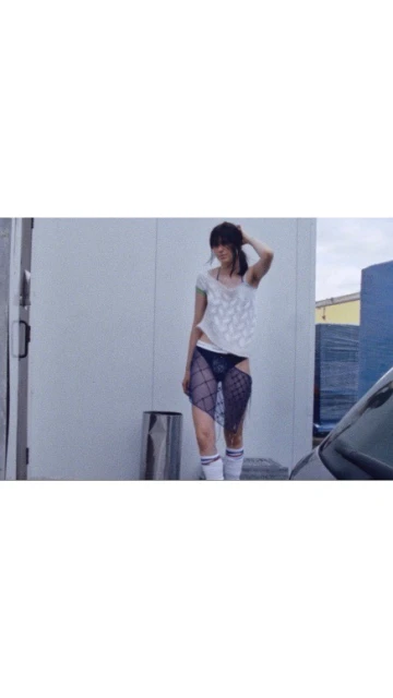 Woman in a white top and fishnet tights poses against an outdoor wall near a metal trash can and parked car.
