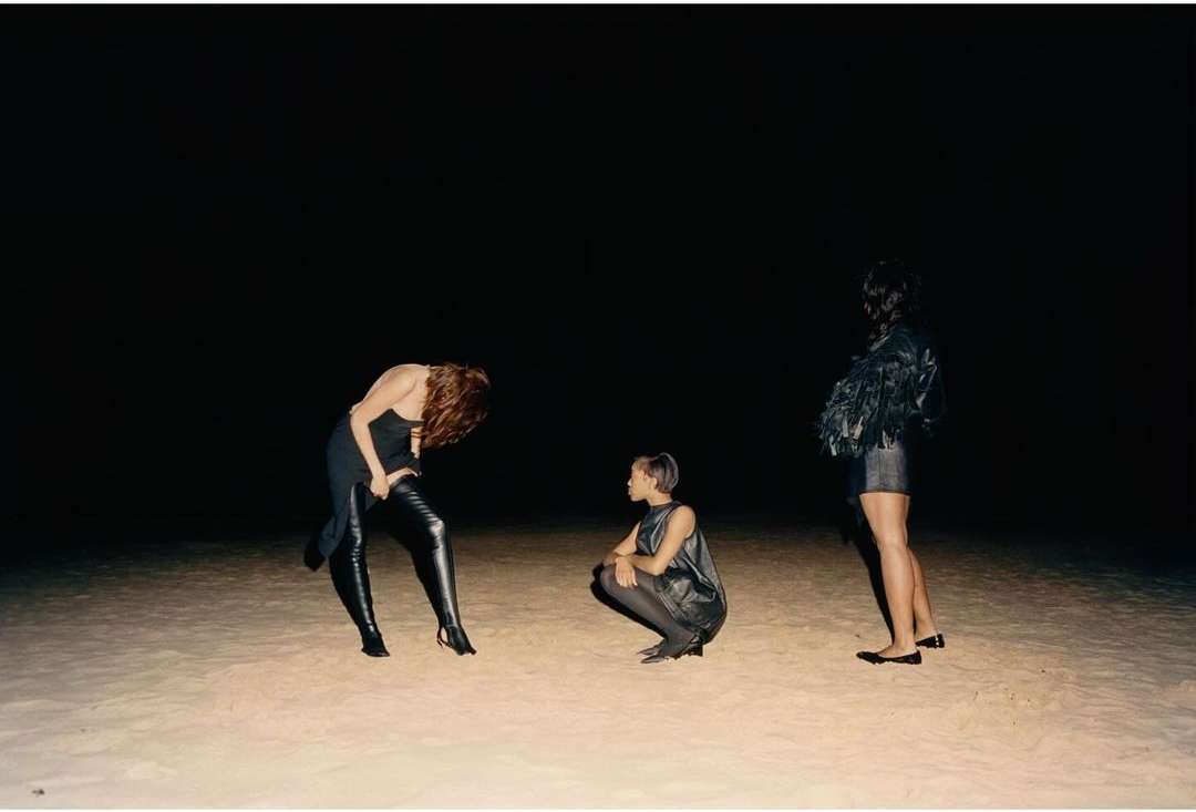 Three women in dark dresses pose on a sandy surface against a black background, one standing, one crouching, and one bent over.