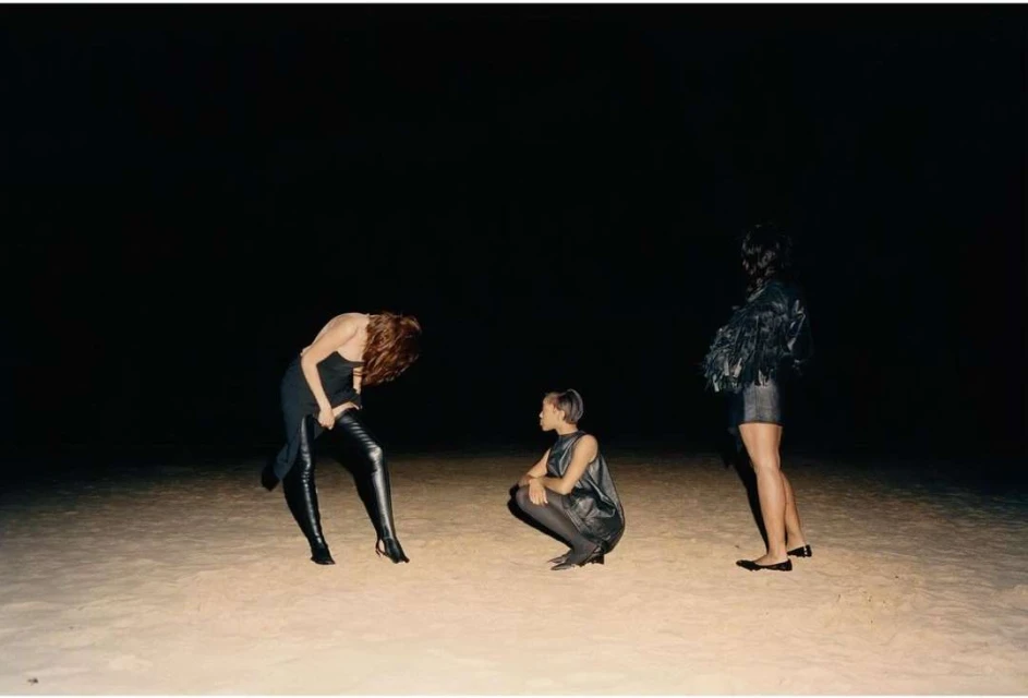Three women in dark dresses pose on a sandy surface against a black background, one standing, one crouching, and one bent over.