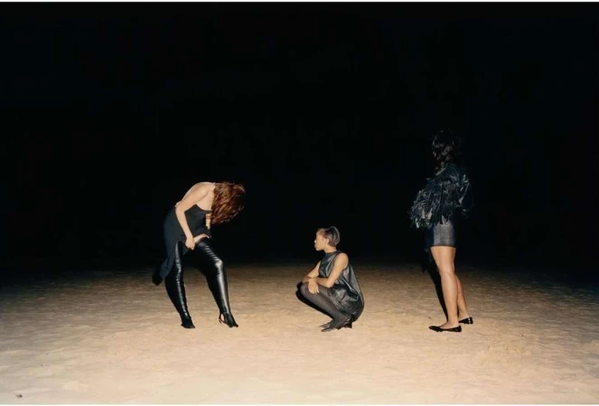 Three women in dark dresses pose on a sandy surface against a black background, one standing, one crouching, and one bent over.