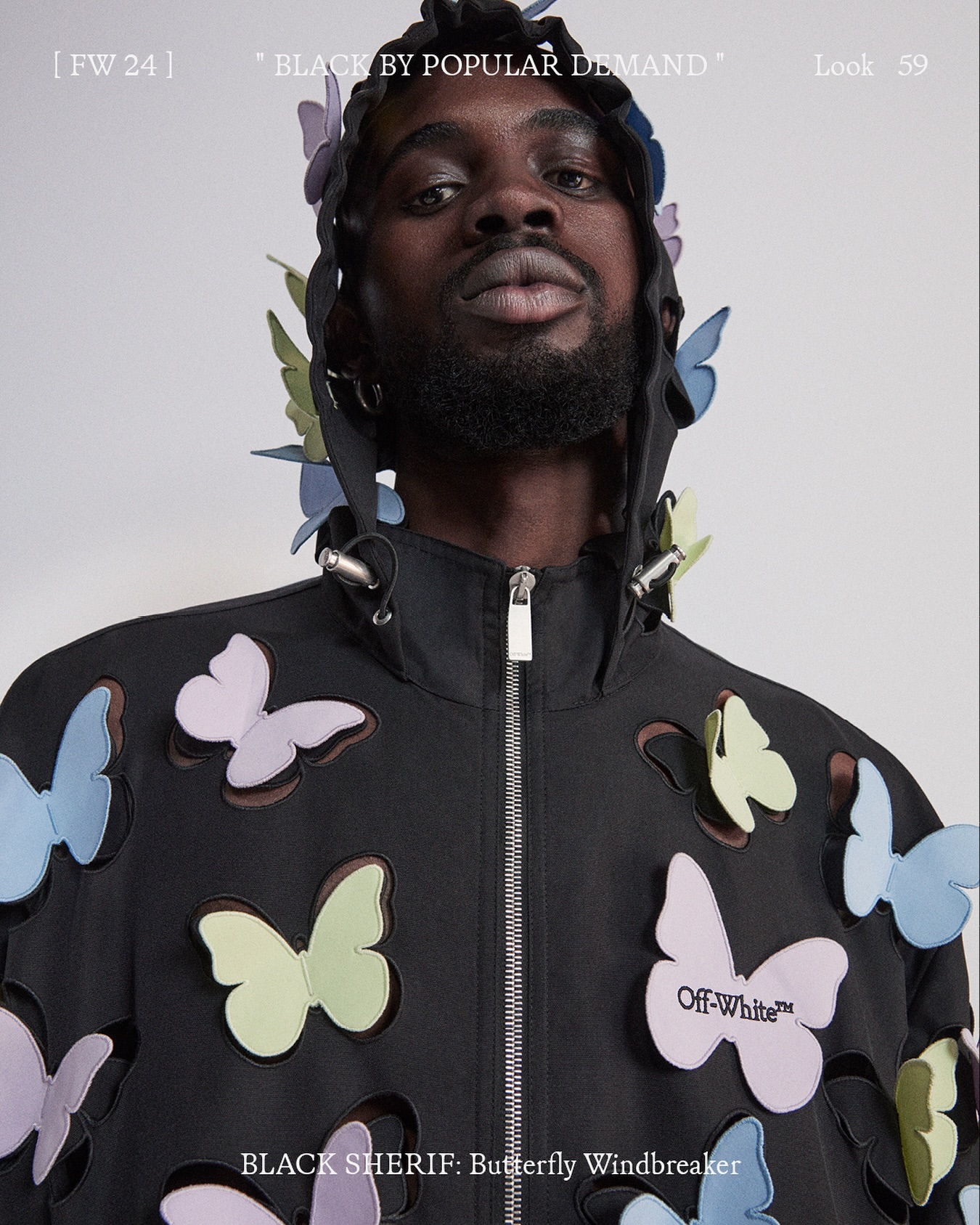 Black man wearing a black windbreaker embellished with multicolored pastel butterfly cutouts against a white background.
