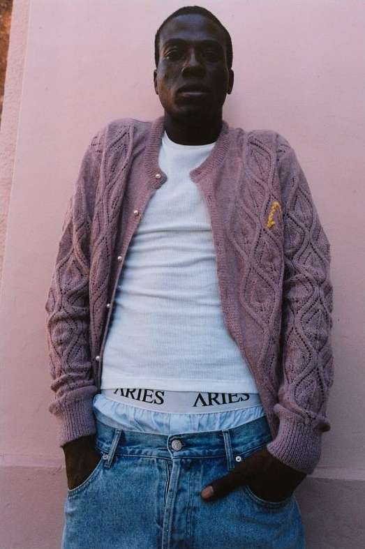 Adult male poses against a light pink wall wearing a lilac cardigan, white t-shirt and light blue jeans with visible underwear band.