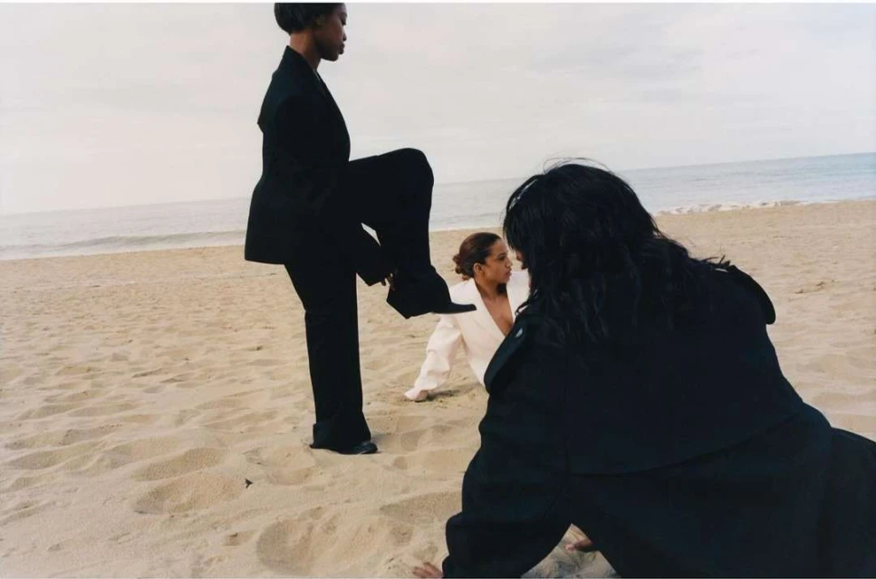 Three adults pose in black and white clothing on a sandy beach with a cloudy sky and ocean backdrop, creating a minimalist fashion scene.