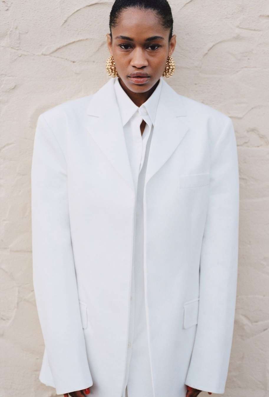 A fashionable Black woman in a white suit with gold earrings poses in front of a textured wall, showcasing modern, elegant style.