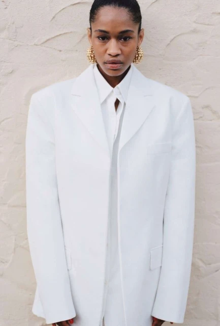 A fashionable Black woman in a white suit with gold earrings poses in front of a textured wall, showcasing modern, elegant style.