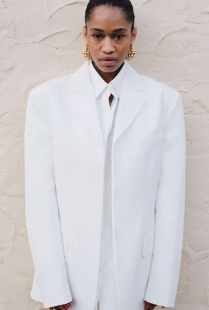 A fashionable Black woman in a white suit with gold earrings poses in front of a textured wall, showcasing modern, elegant style.