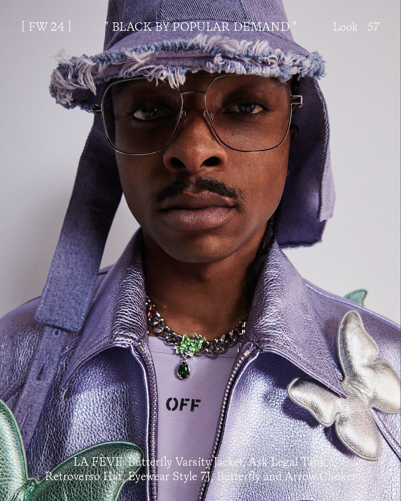 Close-up studio portrait of a Black man in a lavender jacket and hat, glasses, and jewelry, looking directly at the camera.