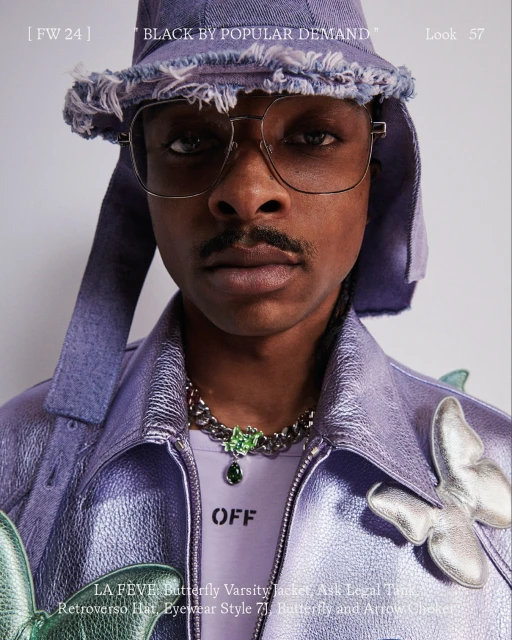 Close-up studio portrait of a Black man in a lavender jacket and hat, glasses, and jewelry, looking directly at the camera.