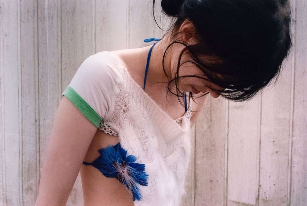 Indoor portrait of a woman with dark hair and blue flower on her arm wearing a white top against a faded white wooden panel background.