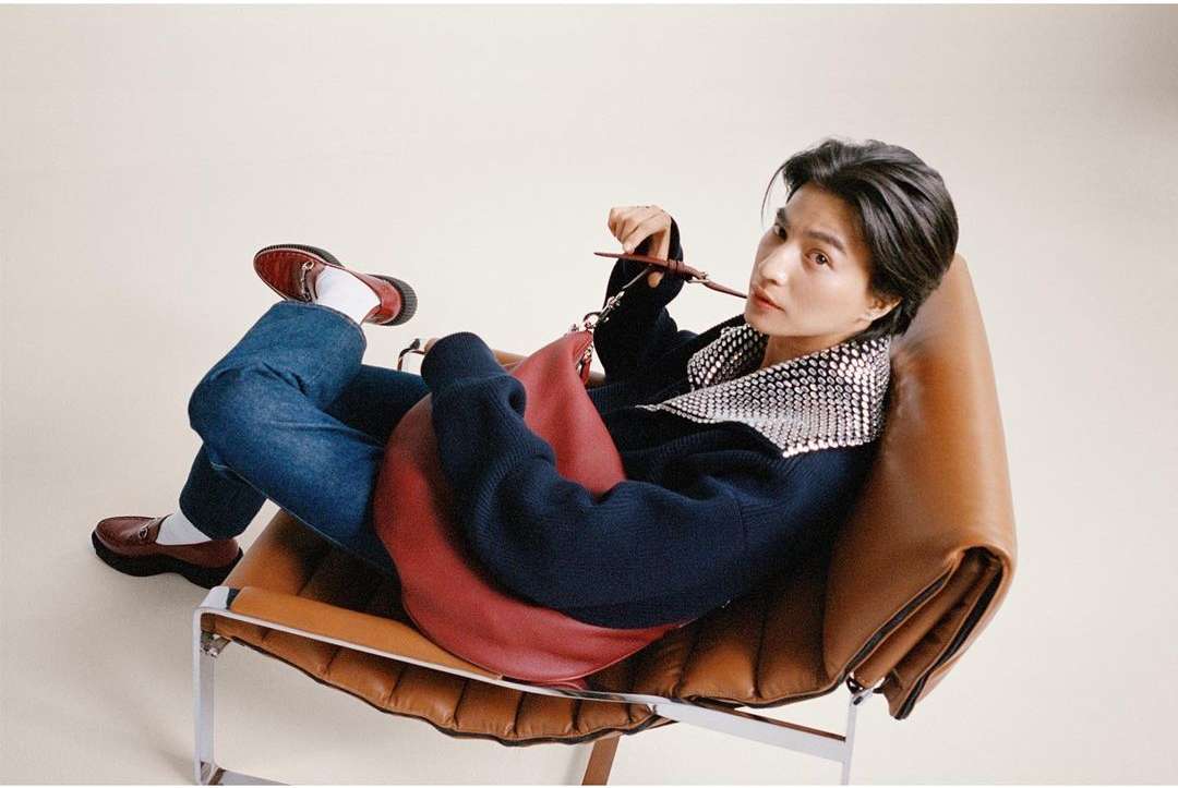 A stylish person lounges in a modern chair, wearing a navy sweater with a jeweled collar, jeans, and loafers with a red handbag nearby.