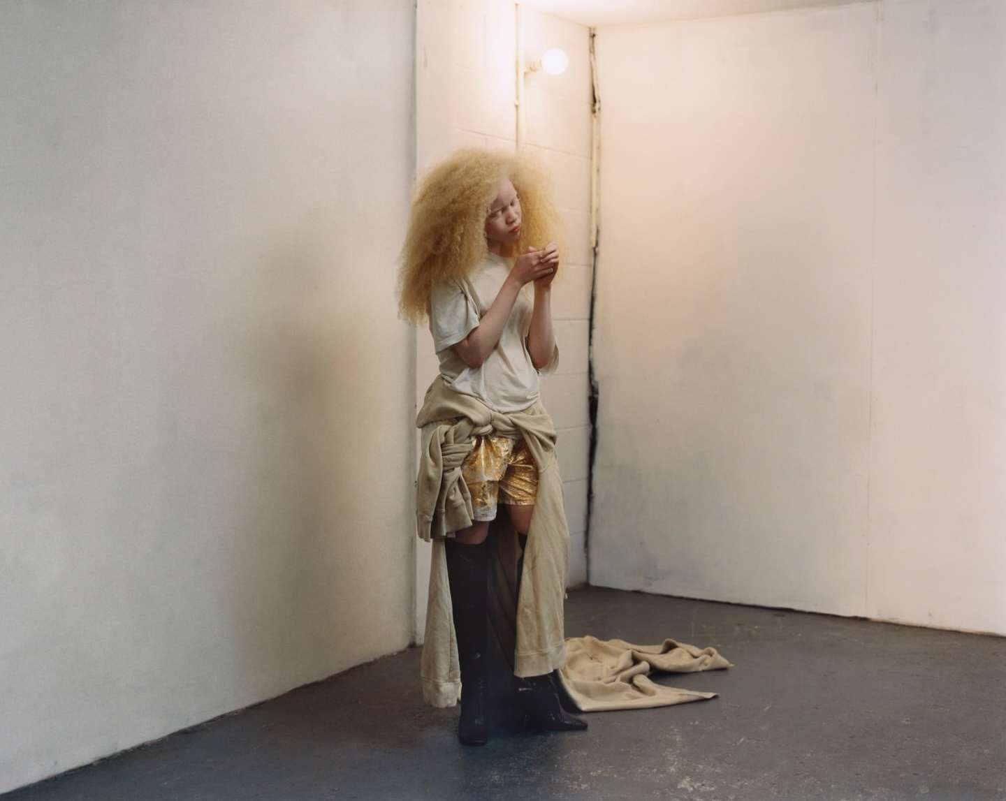 Blonde woman with gold shorts stands in a concrete room with off-white walls, soft light, and dramatic shadows enhancing her distinctive style.