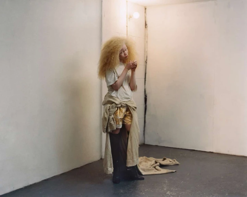Blonde woman with gold shorts stands in a concrete room with off-white walls, soft light, and dramatic shadows enhancing her distinctive style.
