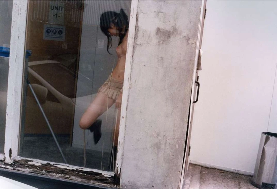 Indoor shot of a young woman in a short skirt posing behind a window. The environment includes weathered walls and an aluminum trash bin.