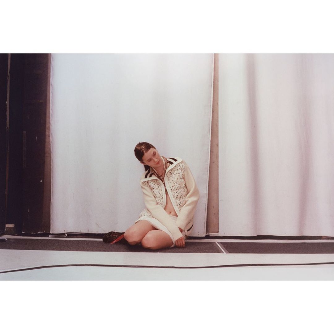 Fashion shot of a fair-skinned adult female in a cream jacket with embellishments kneeling on a dark floor against a white backdrop.