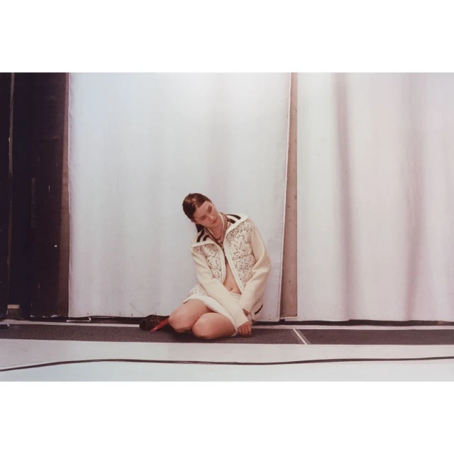 Fashion shot of a fair-skinned adult female in a cream jacket with embellishments kneeling on a dark floor against a white backdrop.