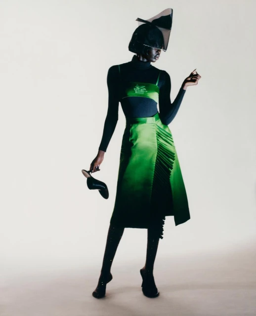 A Black fashion model poses in a green skirt, black top, heels, and a geometric hat against a white backdrop in a studio setting.