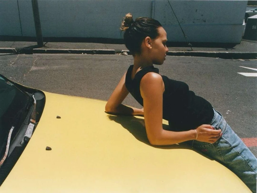 Woman with bun and black tank top leans against the hood of a yellow car parked on a city street on a sunny day.
