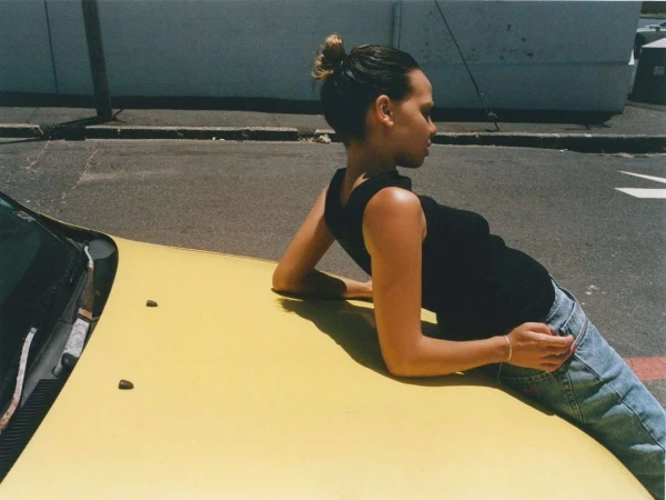 Woman with bun and black tank top leans against the hood of a yellow car parked on a city street on a sunny day.