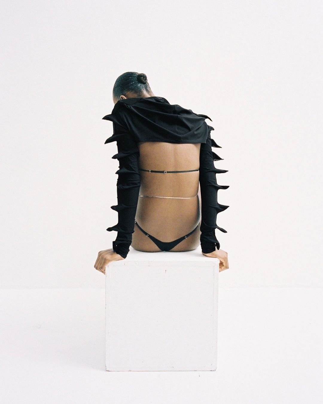 A person with a black hooded top and black thong sits on a white cube, looking down in a minimalistic setting with a bright white background.