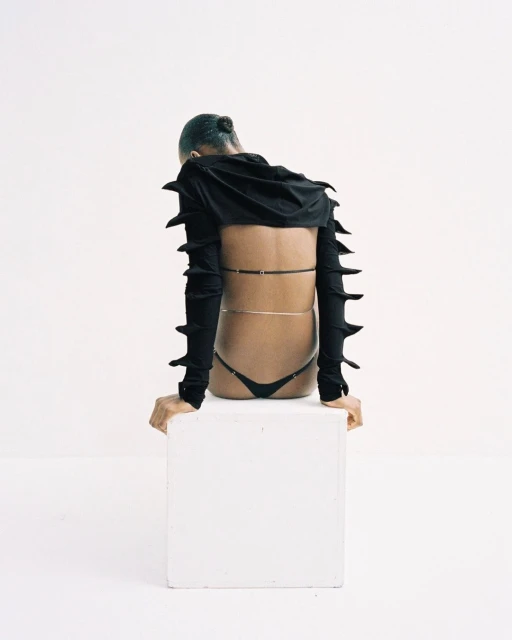 A person with a black hooded top and black thong sits on a white cube, looking down in a minimalistic setting with a bright white background.