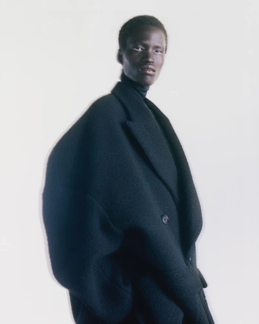 Fashion portrait of a dark-skinned female model wearing a black turtleneck and oversized wool coat against a bright, soft white background.