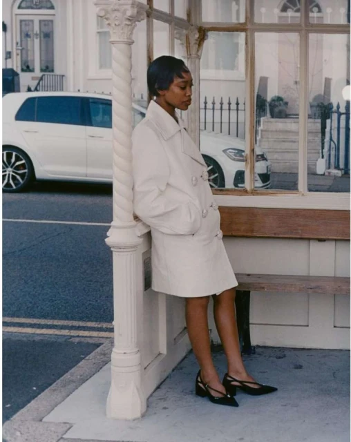 An elegant Black woman in a white coat leans against a bus stop in an urban setting, with parked cars visible, showcasing fashion and style.