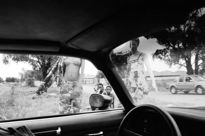 Black and white photograph looking out through a car window at several people standing outside by a street.