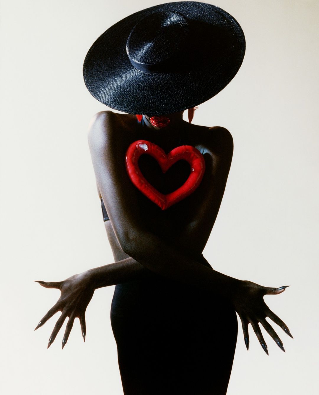 Elegant dark-skinned woman with dramatic black hat, long black nails, and a painted heart on her back, posing against a pale backdrop.