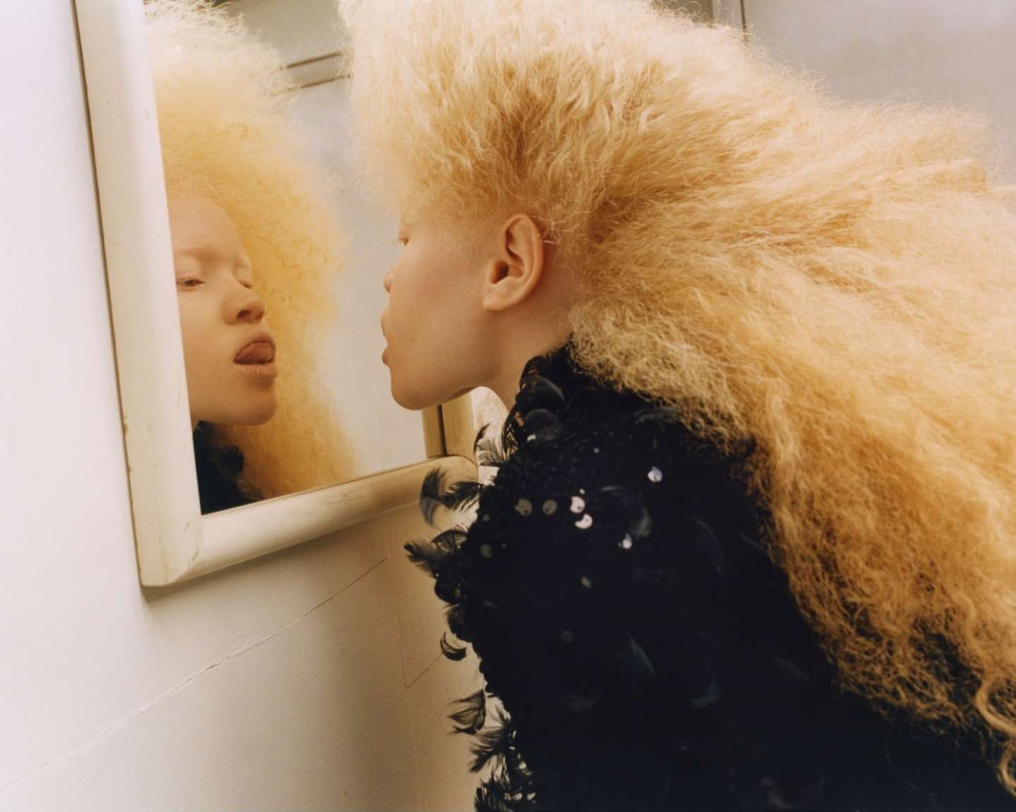 Woman with blonde afro hairstyle poses near mirror, wearing black dress, indoors with bright lighting, showing tongue to reflection.