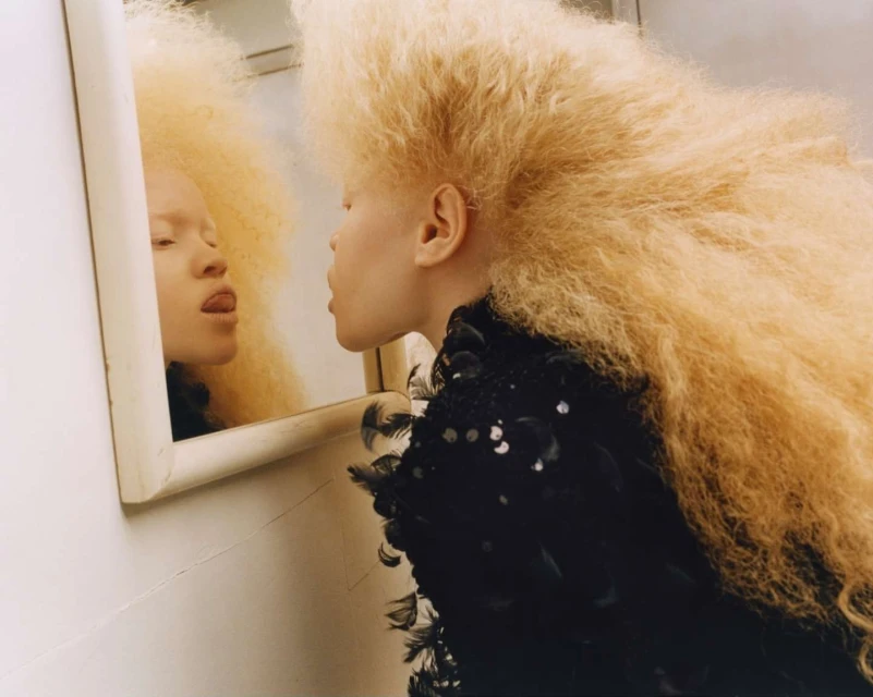 Woman with blonde afro hairstyle poses near mirror, wearing black dress, indoors with bright lighting, showing tongue to reflection.