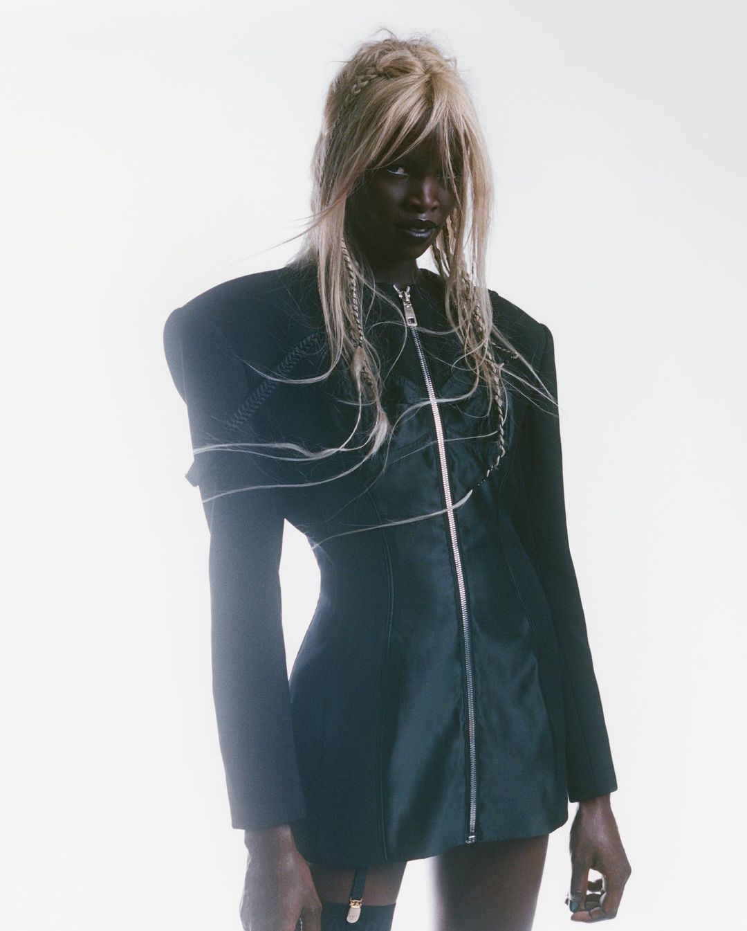 Fashion portrait of a slender, dark-skinned woman with blond hair, wearing a black mini dress with a zipper and long sleeves against a white background.
