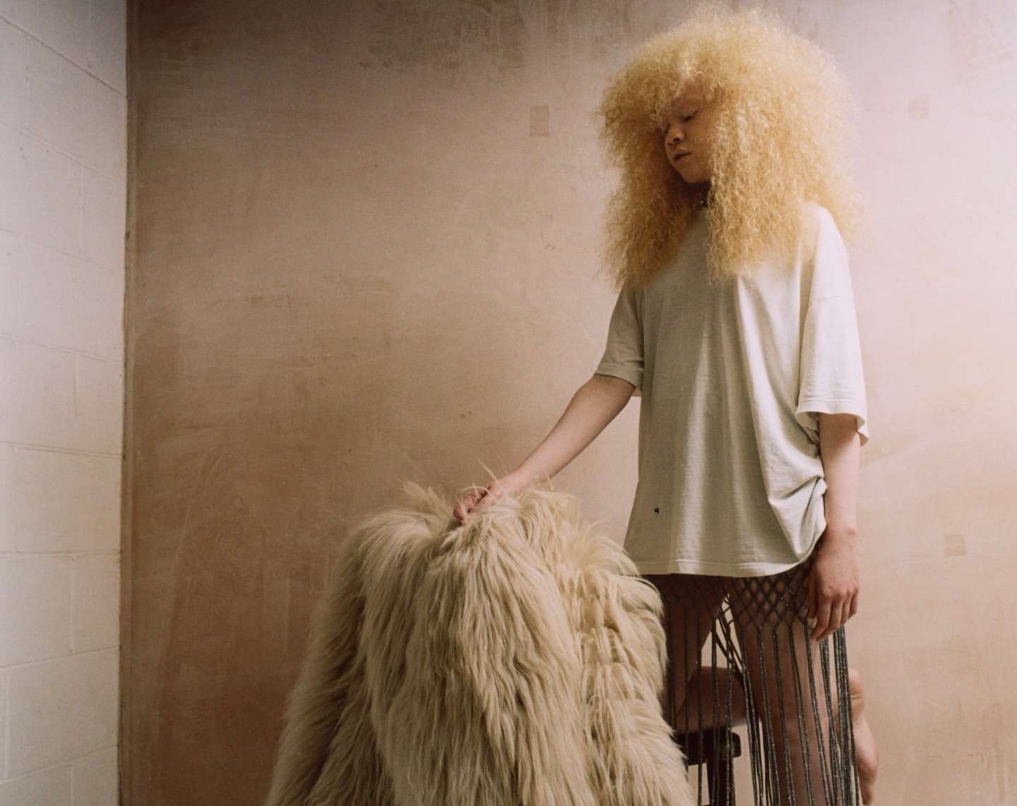 Woman with blonde afro and fringed skirt touches a shaggy coat in front of a light-brown plaster wall and white tiled wall.