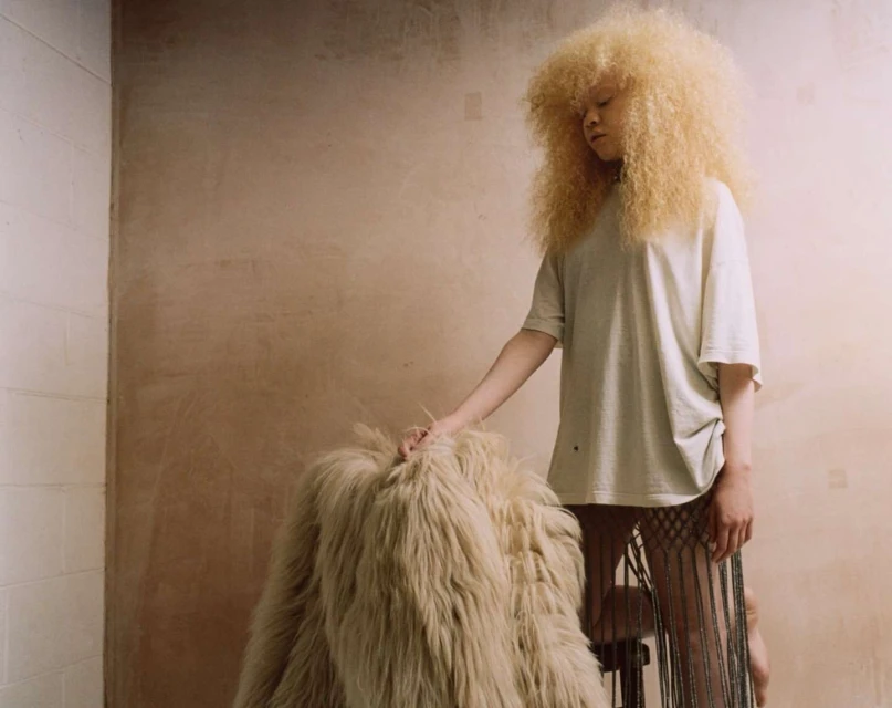 Woman with blonde afro and fringed skirt touches a shaggy coat in front of a light-brown plaster wall and white tiled wall.