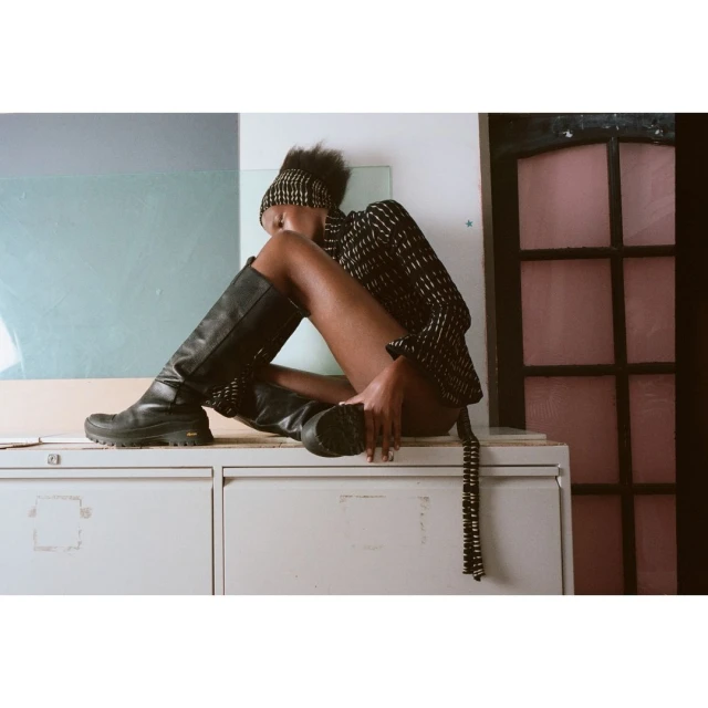 A woman poses in a patterned top, black boots, and a headband while sitting on a filing cabinet in a brightly lit indoor setting.