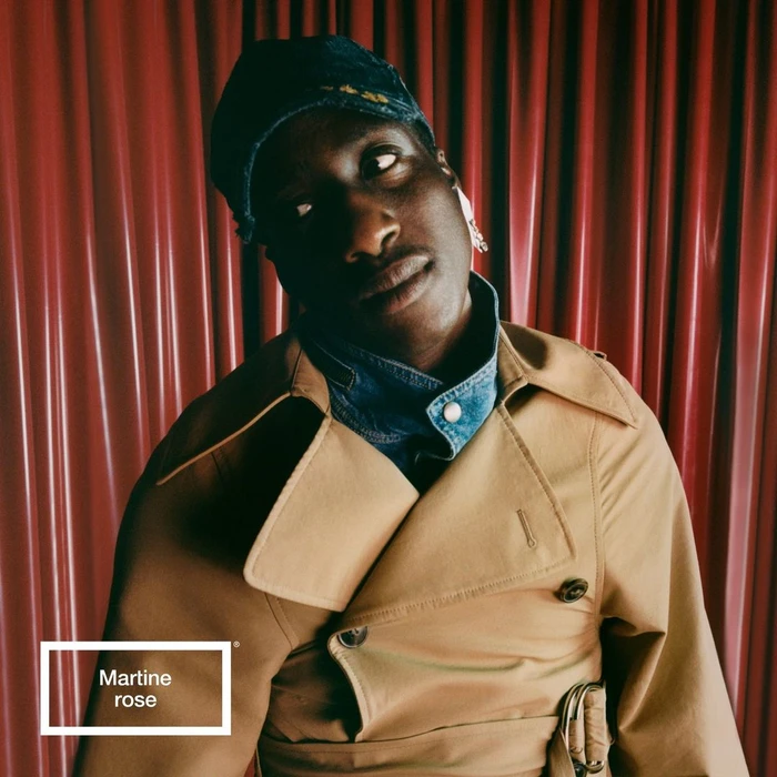 Portrait of a black man wearing a tan trench coat, denim jacket collar, and blue cap against a red curtain background.