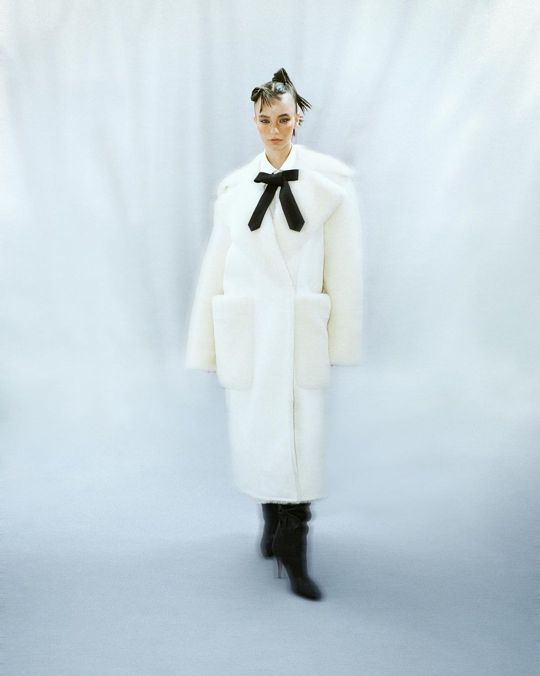 Model in a long white faux fur coat with a black bow and dark boots standing against a draped white background.