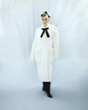 Model in a long white faux fur coat with a black bow and dark boots standing against a draped white background.