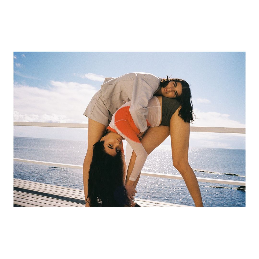 Two women in sportswear pose acrobatically on a deck overlooking the ocean under a clear blue sky with minimal clouds, captured in daylight.