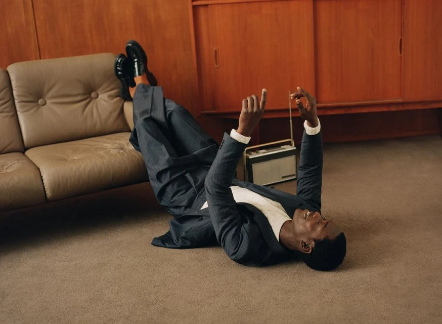 Young man in a suit laying on a carpeted floor near a leather sofa, smiling with his hands raised, wearing black dress shoes next to a radio.