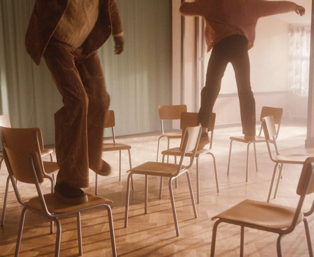 Two people jumping across chairs in a room with light streaming through a window. Chairs are brown and silver.