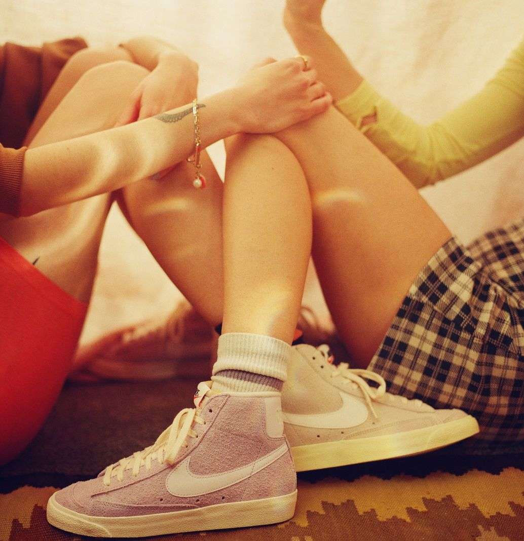 Two people wearing vintage Nike Blazer sneakers sitting indoors, one in lilac and the other in white, casual lifestyle fashion shoot.