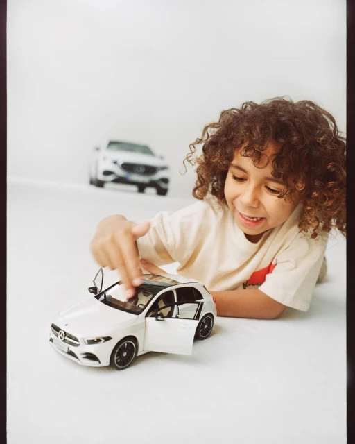 A child plays with a toy car, showcasing its open doors, with another car blurred in the background against a bright studio backdrop.