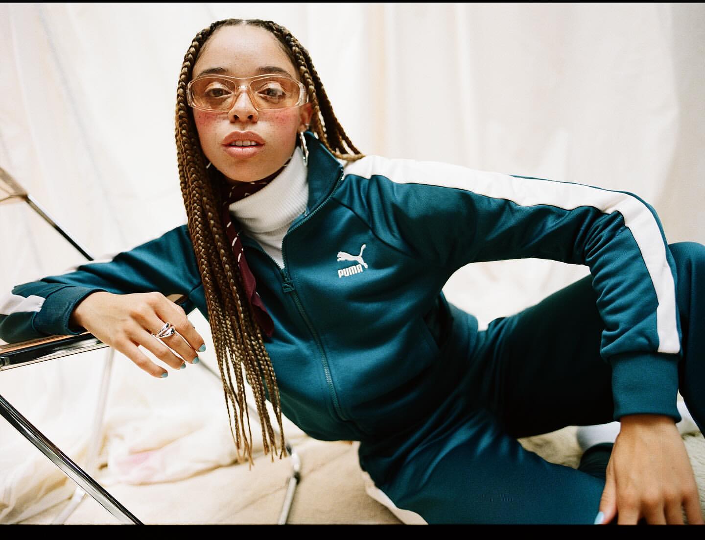 A woman in a teal Puma tracksuit poses indoors, wearing tinted glasses, with braided hair and light blue nail polish.
