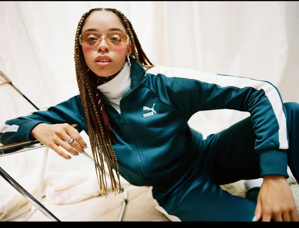 A woman in a teal Puma tracksuit poses indoors, wearing tinted glasses, with braided hair and light blue nail polish.