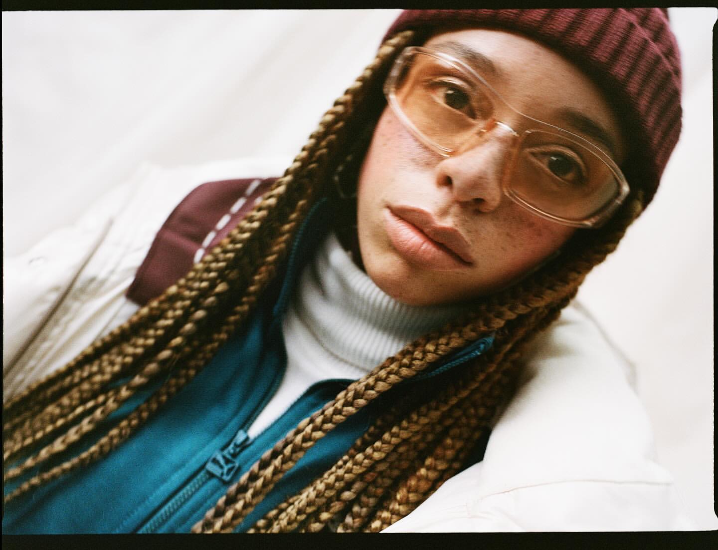 Portrait of an adult woman with braided hair, wearing a beanie hat and retro glasses in a close-up studio shot.