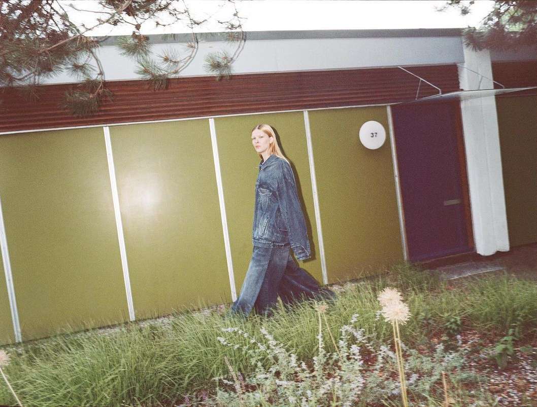 A blonde female fashion model in a denim outfit walks in front of a modern, olive green residential home with a blue door.