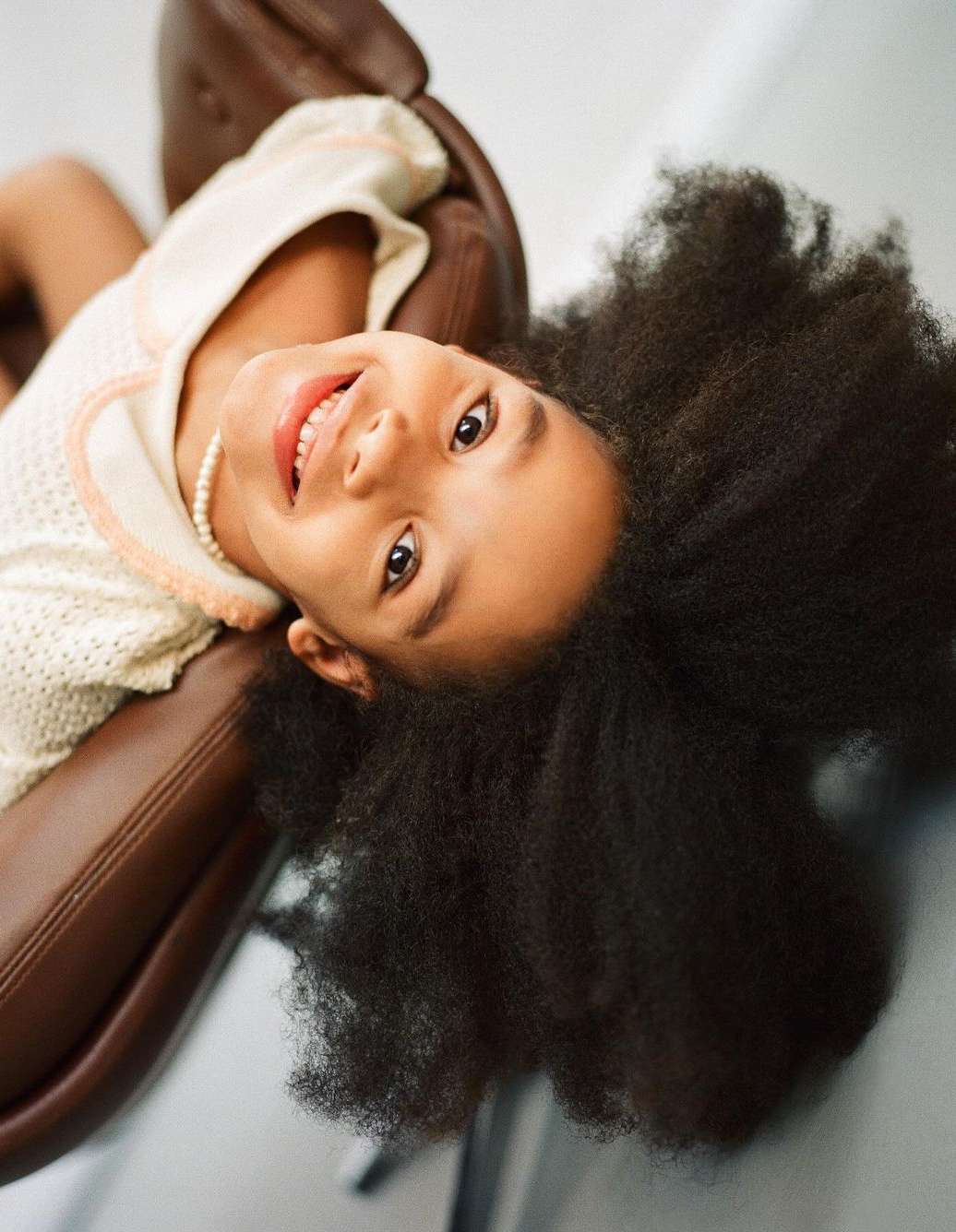A young Black child with voluminous dark curly hair smiling upside down in a brown leather chair, captured by Jessica Sidenros.