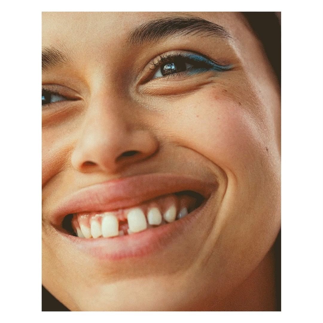 Close-up portrait of a smiling young adult woman with fair skin, blue eyeliner, and a gap between her front teeth, showcasing a happy expression.